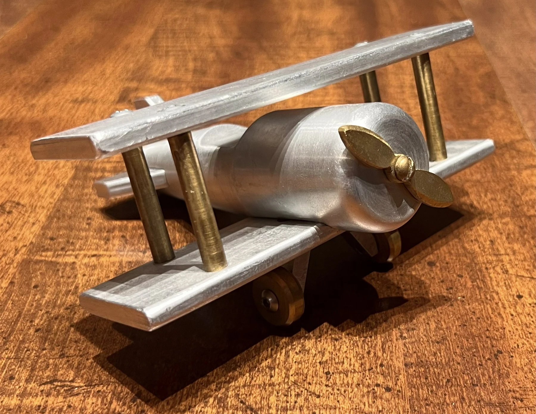 The finished aluminum and brass model plane on a wooden table, showing the machined wings, propeller, and wheels.