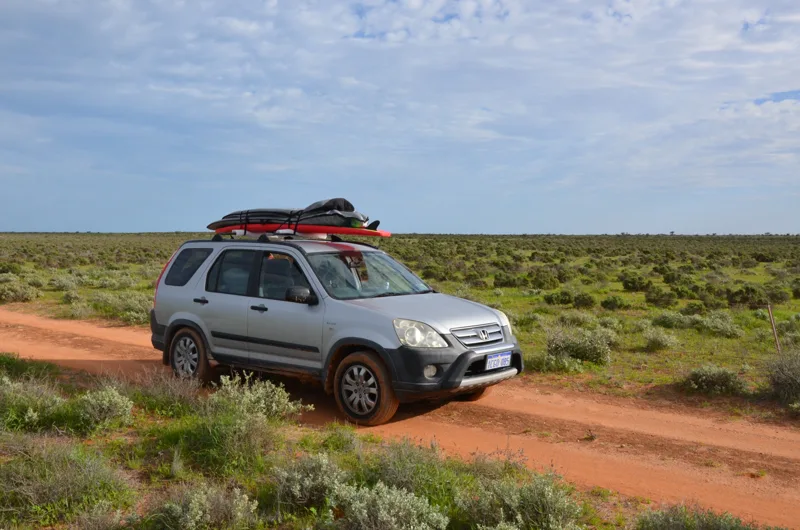 A silver vehicle carrying a kayak along a red dirt track.